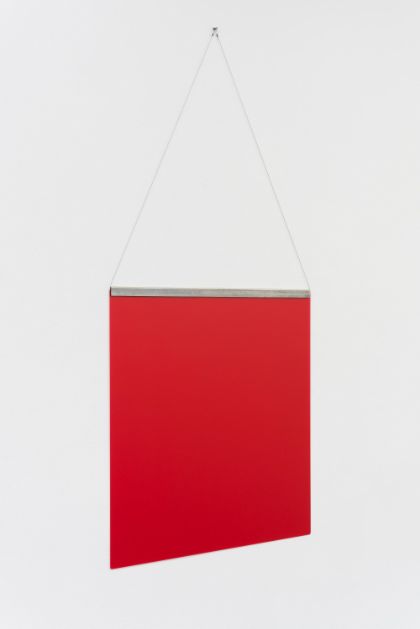 A thin, square red surface hangs on a white wall, attached along its upper edge by taut threads connected to a narrow metal frame. The photograph shows the object from a side perspective.
