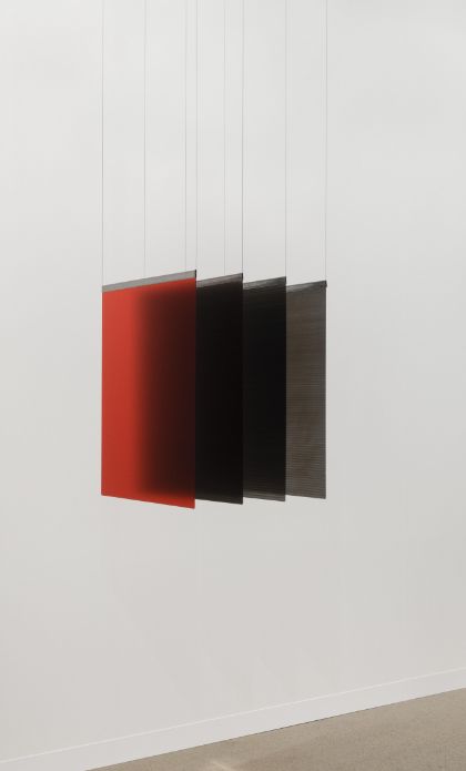 Four equally sized, semi-transparent squares are arranged parallel to one another in front of a white wall. The front square is red, followed by squares in black, grey, and brown tones. All squares are suspended from the ceiling by fine threads attached at their upper corners. The photograph shows the artwork from a side perspective.