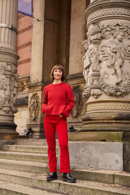 Jenny Schlenzka steht lächelnd in einem roten Kostüm auf den Stufen zum Eingang des Gropius Bau.