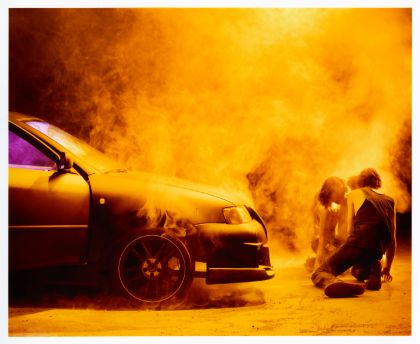 Zwei Performer*innen knien auf der Bühne vor einem Auto, um sie herum ist es nebelig.